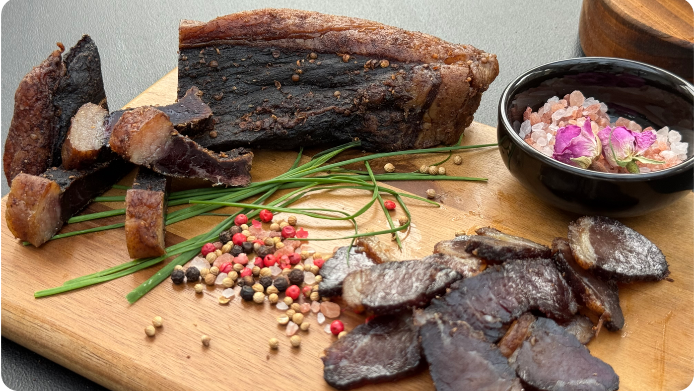 Dried Biltong, whole, chunked and slices from Biltong Direct on a wooden board with spices and a small bowl of pink flowers.