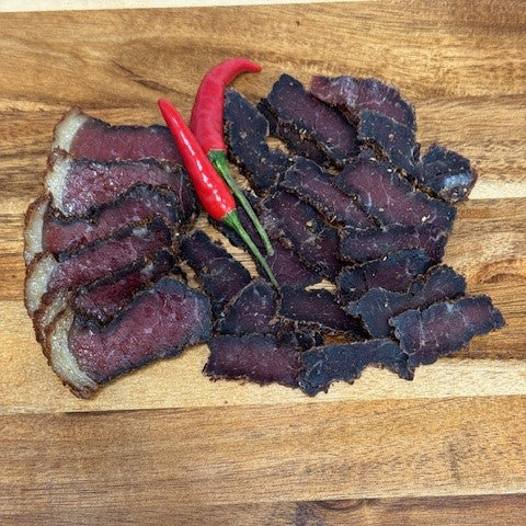 Sliced biltong from Biltong Direct arranged on a wooden surface with a red chili pepper.