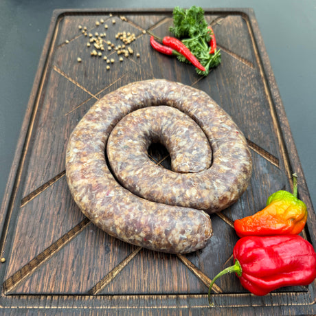 Coil of raw chilli boerewors sausage on a wooden cutting board with chilli peppers and herbs