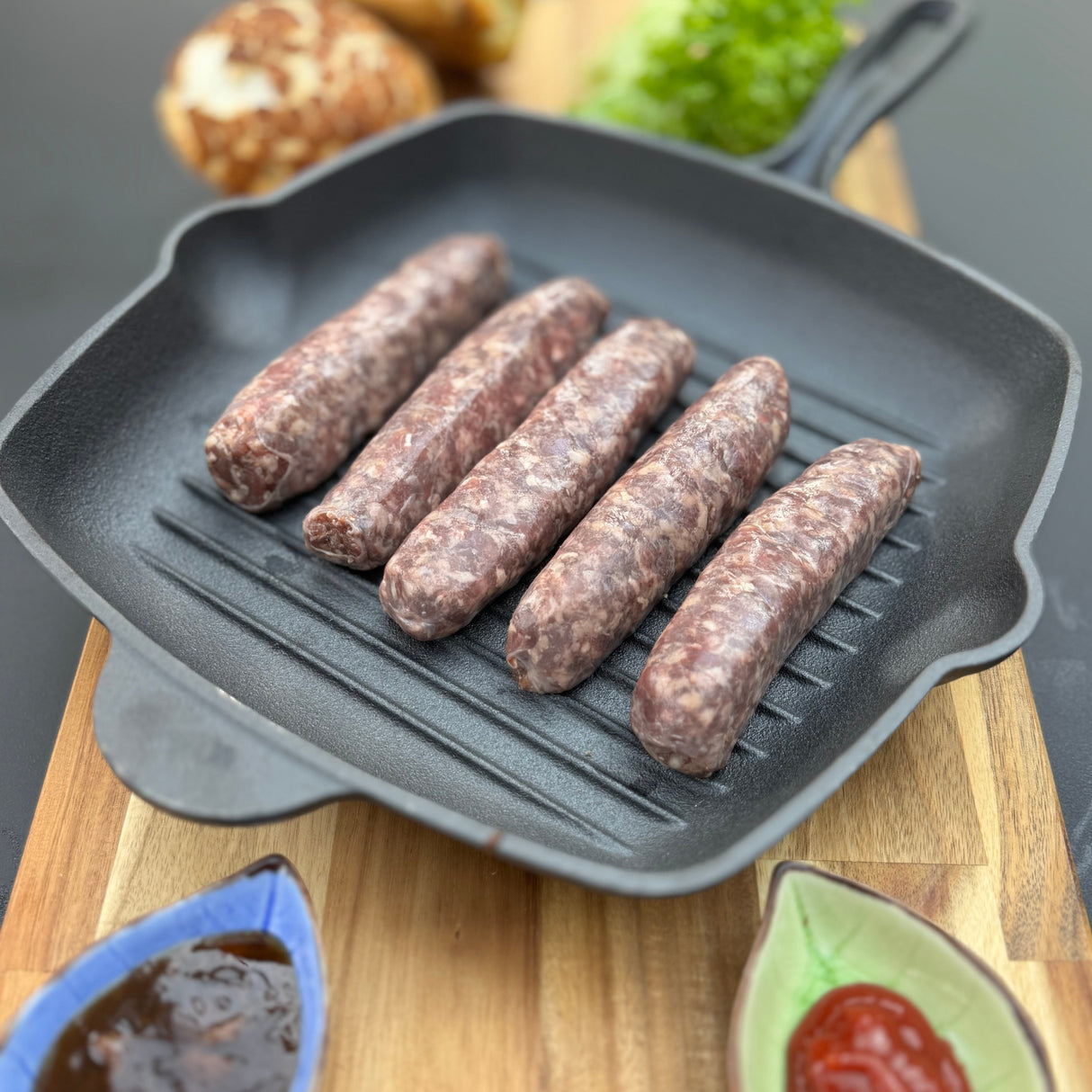 Raw Traditional boerewors sausages from Biltong Direct in a grill pan on a wooden cutting board with ingredients in the background.