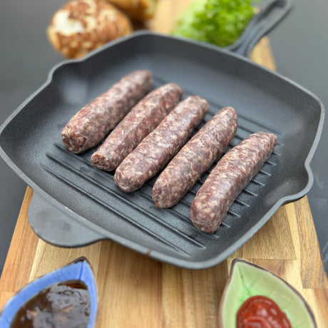 Raw Traditional boerewors sausages from Biltong Direct in a grill pan on a wooden cutting board with ingredients in the background.