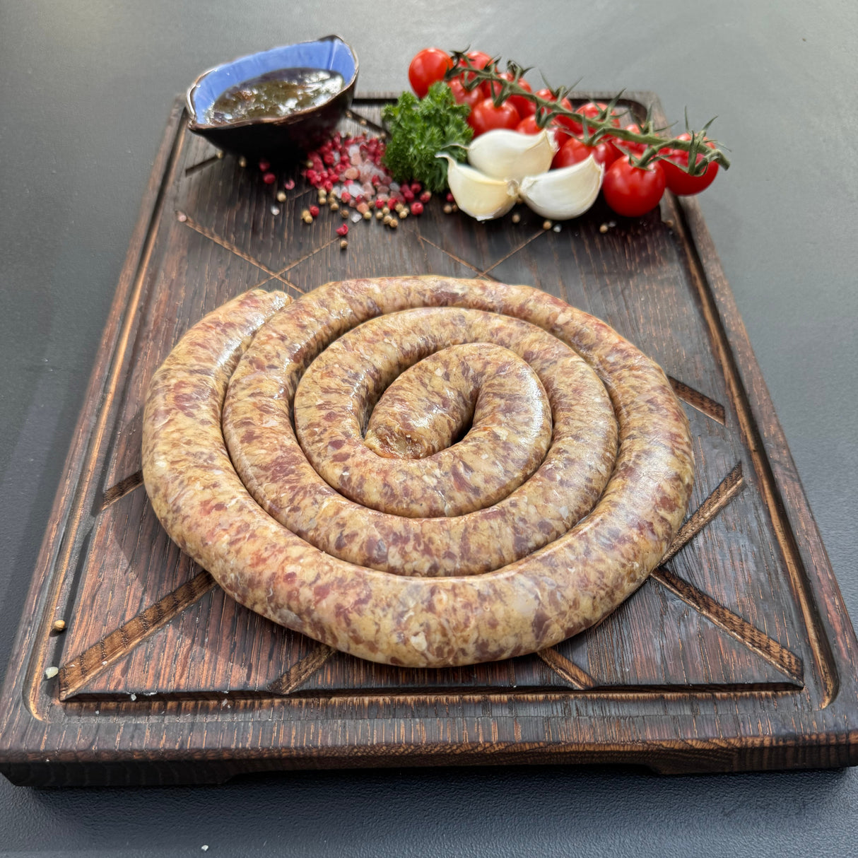 Coiled raw sosatie mini boerewors sausage from Biltong Direct on a wooden cutting board with tomatoes and garlic and Mrs Balls Chutney.
