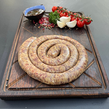 Coiled raw sosatie mini boerewors sausage from Biltong Direct on a wooden cutting board with tomatoes and garlic and Mrs Balls Chutney.