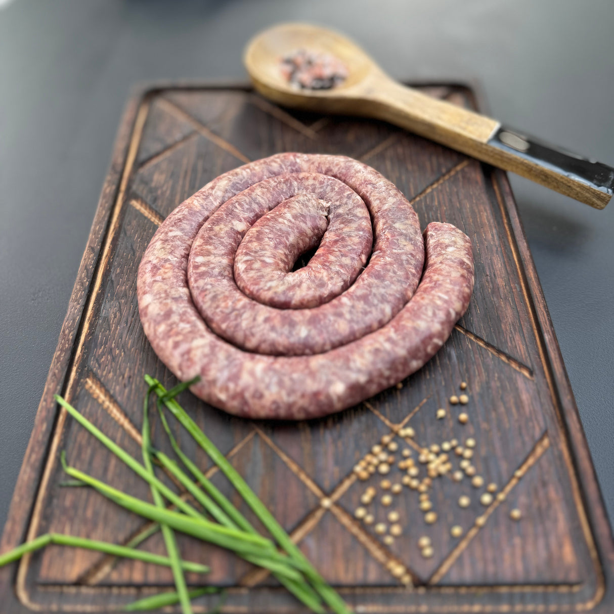 Coiled raw traditional mini boerewors sausage from Biltong Direct on a wooden cutting board with chives and mustard seeds.