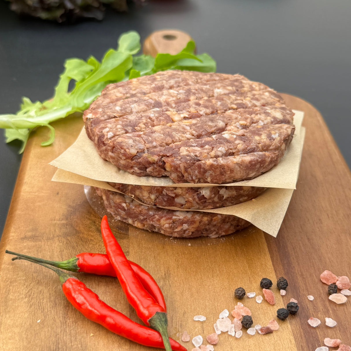 A stack of raw Chilli boere burger patties from Biltong Direct on a wooden cutting board with red peppers and spices.