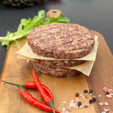 A stack of raw Chilli  boere burger patties from Biltong Direct on a wooden cutting board with red peppers and spices.