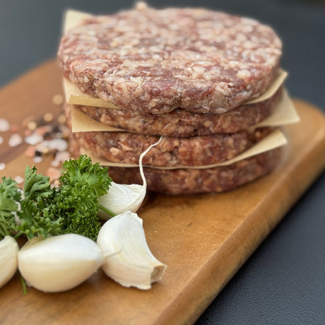 A stack of raw garlic boere burger patties from Biltong Direct on a wooden cutting board with herbs and garlic.