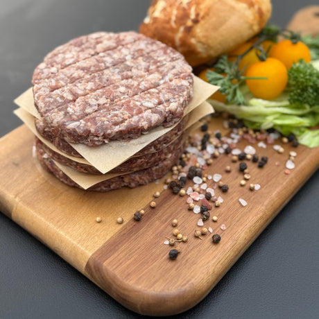 A stack of raw traditional boere burger patties from Biltong Direct on a wooden board with bread and vegetables in the background
