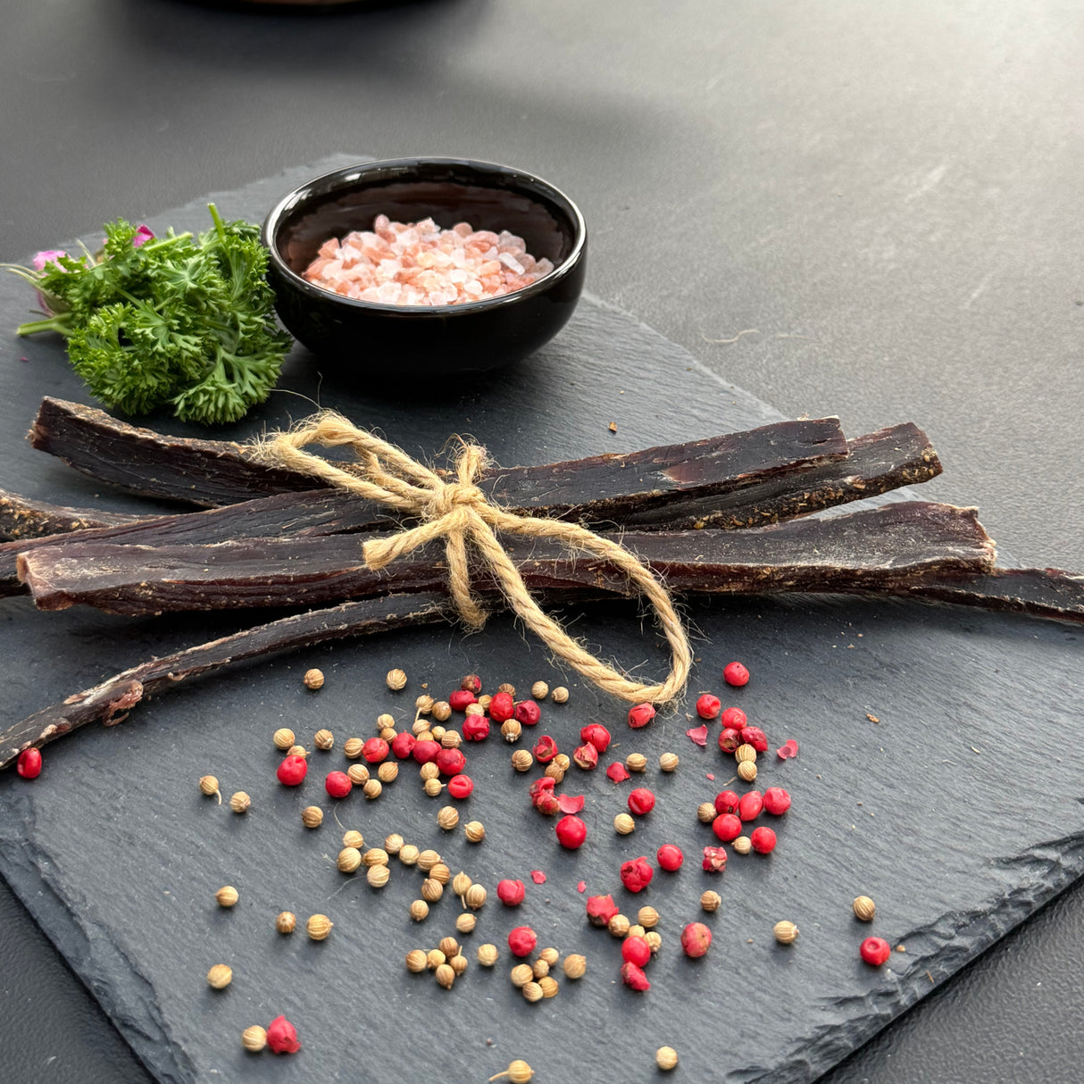 Dried beef biltong sticks from Biltong Direct on a slate surface with spices and a bowl of pink salt.