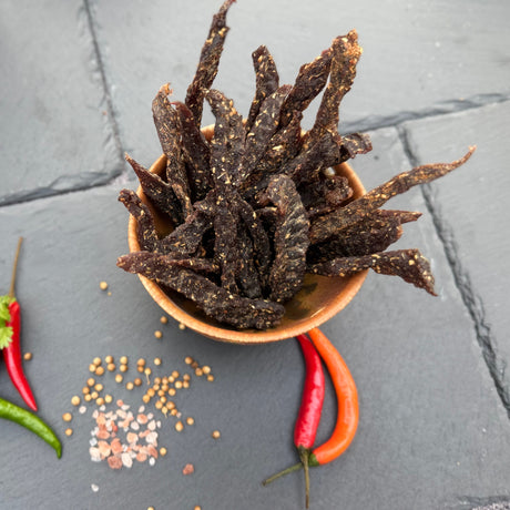 Dried beef biltong snap sticks or stokkies from Biltong Direct in a bowl on a grey surface with red and green peppers and spices.
