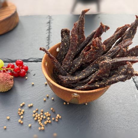 Dried beef biltong snap sticks or stokkies from Biltong Direct in a terracotta bowl on a dark surface with spices and fruits.