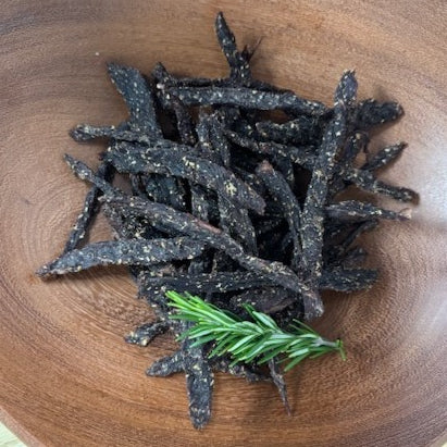 A wooden bowl filled with air-dried, gluten-free beef snap sticks, also known as Stokkies, with a sprig of rosemary to the side.