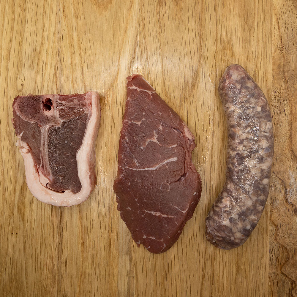 Assorted raw meats on a wooden surface, including a piece of boerewors, rump steak, and a lamb chop from Biltong Direct.