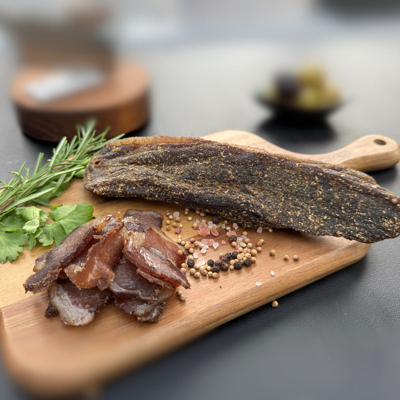 Dried beef biltong a whole piece and slices from Biltong Direct Biltong on a wooden board with herbs and spices.