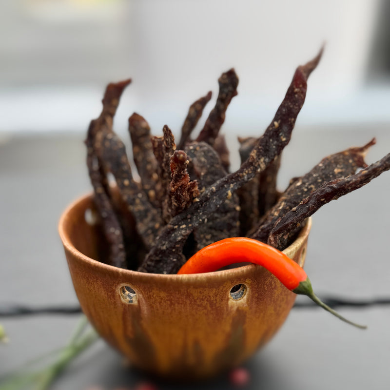 Dried beef snap sticks or stokkies  from Biltong Direct in a small brown bowl with a red chili pepper on a blurred background