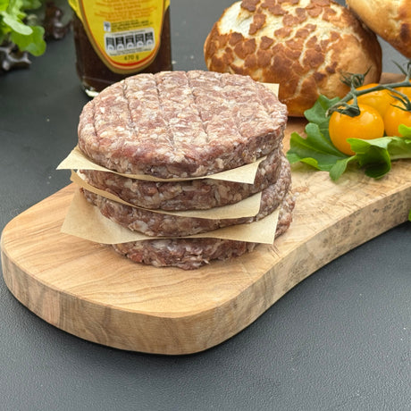 A stack of raw gluten free boere burger patties from Biltong Direct on a wooden cutting board with bread and vegetables in the background.