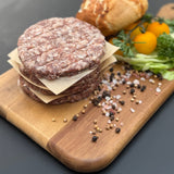 A stack of raw traditional boere burger patties from Biltong Direct on a wooden board with bread and vegetables in the background