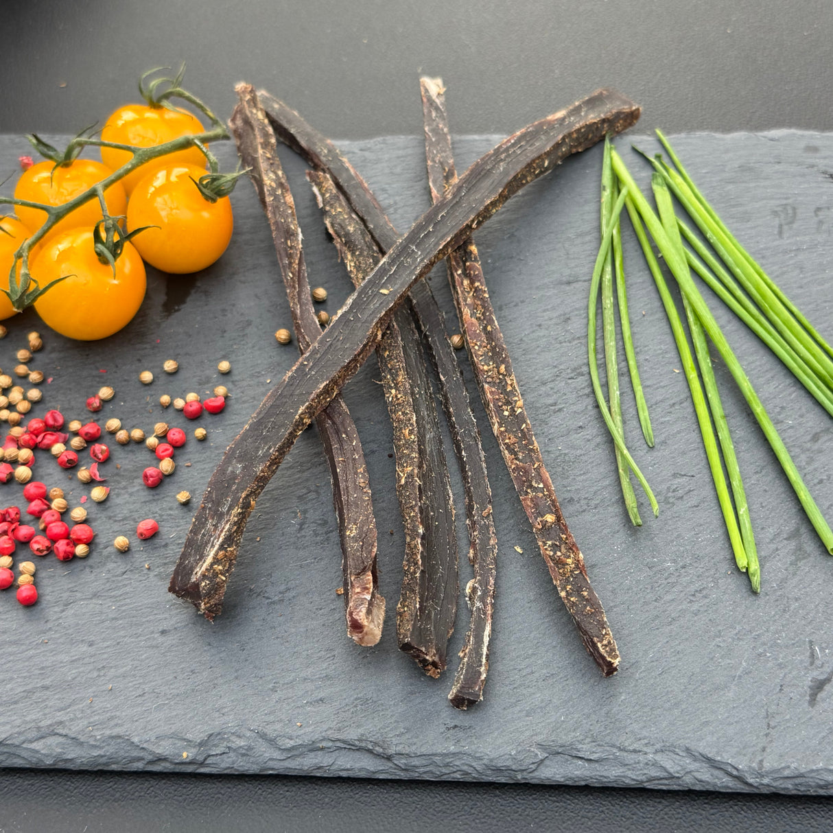 Dried beef biltong strips from Biltong Direct on a slate surface with yellow tomatoes, green chives, and colourful peppercorns.