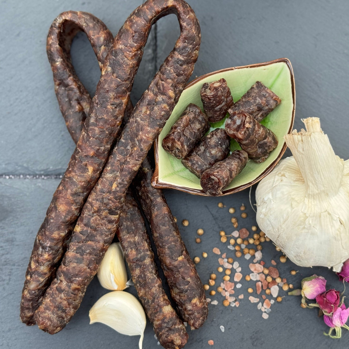 Garlic Droewors or drywors, a dried sausage from Biltong Direct with a small bowl of smaller pieces, garlic, and spices on a dark surface.