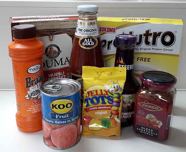Assorted food items including a can of Koo fruit, a bottle of All Gold tomato sauce, Braai salt, and a box of cereal on a white surface.